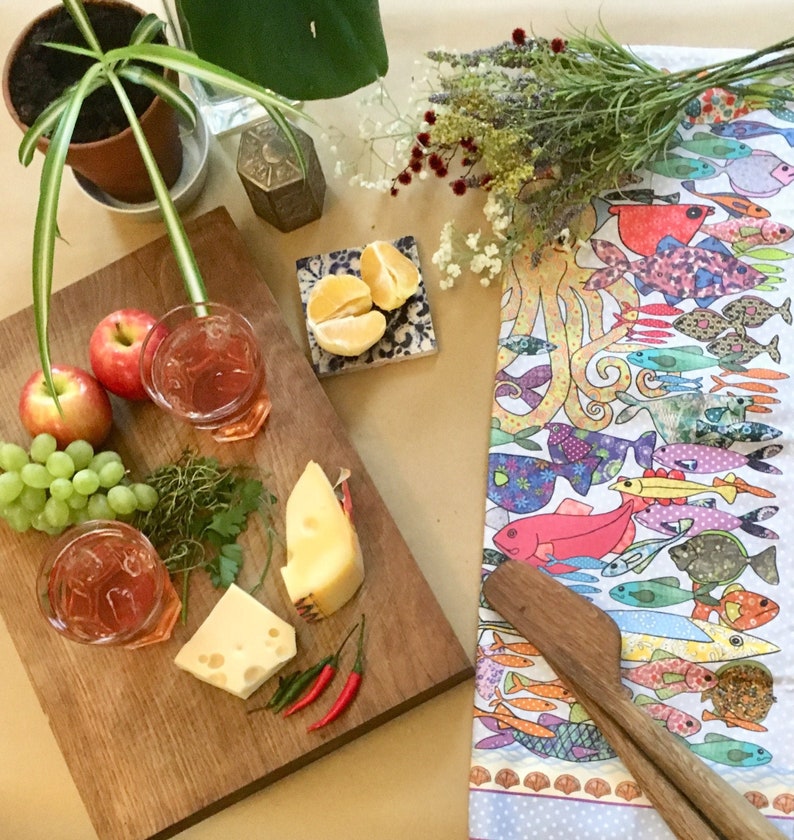 May include: A wooden cutting board with a variety of snacks, including fruit, cheese, and drinks. The board is set on a table with a colorful tablecloth featuring a pattern of fish and an octopus.