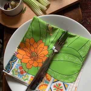 Puede incluir: Un plato blanco con una servilleta floral verde y naranja doblada y un tenedor plateado. La servilleta presenta una gran flor naranja y un peque&ntilde;o dise&ntilde;o de rana. Aceitunas, apio y un vaso de zumo en el fondo.