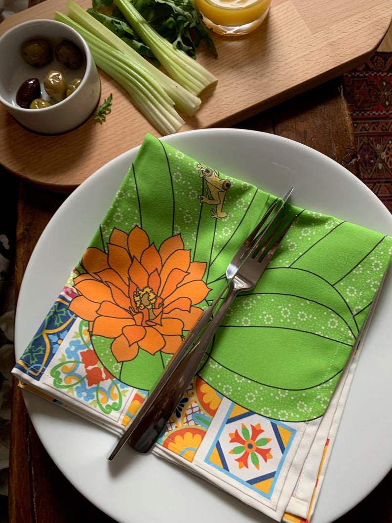 Puede incluir: Un plato blanco con una servilleta floral verde y naranja doblada y un tenedor plateado. La servilleta presenta una gran flor naranja y un peque&ntilde;o dise&ntilde;o de rana. Aceitunas, apio y un vaso de zumo en el fondo.