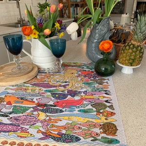 May include: A colorful table runner with a pattern of fish and an octopus. The runner is on a white table with a vase of white and yellow flowers.