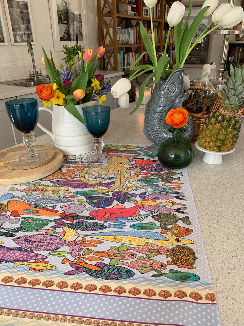 May include: A colorful table runner with a pattern of fish and an octopus. The runner is on a white table with a vase of white and yellow flowers.