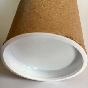 Puede incluir: Un tubo de cart&oacute;n marr&oacute;n con una tapa de pl&aacute;stico blanco. El tubo est&aacute; vac&iacute;o y la tapa es lisa.
