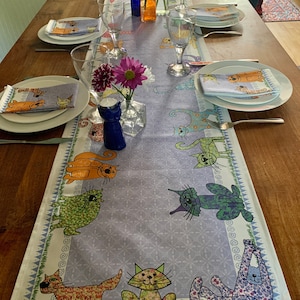 May include: A purple and white table runner featuring a repeating pattern of colourful cats. The runner is laid out on a wooden table with place settings and flowers in a vase.