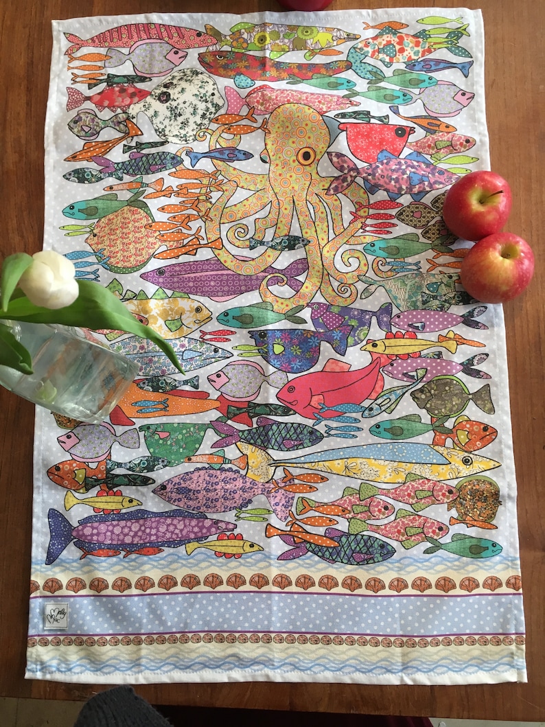 May include: A white tea towel with a colorful design of an octopus surrounded by fish. The fish are all different shapes and sizes and have a variety of patterns. The octopus is yellow with orange circles and has eight tentacles. The tea towel has a blue and white border with a scalloped edge.