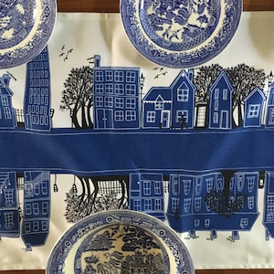 May include: A white table runner featuring a blue and white pattern of houses, trees, and birds. The runner is adorned with blue and white porcelain plates with a floral design.