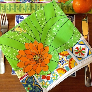 Puede incluir: Una servilleta floral verde y blanca con un dise&ntilde;o de flor naranja y rana amarilla. La servilleta est&aacute; doblada y colocada sobre una mesa con tenedor y cuchillo. La servilleta tiene un borde colorido con patrones geom&eacute;tricos.
