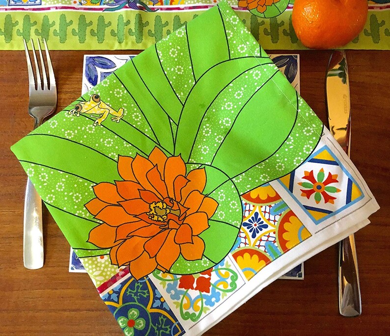 Puede incluir: Una servilleta floral verde y blanca con un dise&ntilde;o de flor naranja y rana amarilla. La servilleta est&aacute; doblada y colocada sobre una mesa con tenedor y cuchillo. La servilleta tiene un borde colorido con patrones geom&eacute;tricos.