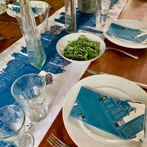 May include: A dining table set with a blue and white cityscape table runner. White plates, blue napkins, and clear glasses are arranged on the wooden table. A bowl of green salad and decorative bottles complete the setting. The table setting is ready for a meal.