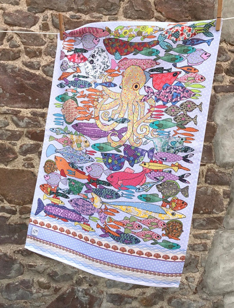May include: A white tea towel with a colorful design of an octopus surrounded by fish. The fish are all different shapes, sizes, and colors. The octopus is yellow with brown spots and has eight tentacles. The tea towel has a blue border with a white polka dot pattern.