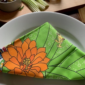 Puede incluir: Una servilleta plegada con estampado floral verde y naranja sobre un plato blanco. La servilleta presenta una gran flor naranja y una rana de dibujos animados. El fondo incluye una tabla de cortar de madera, apio, aceitunas y un vaso de zumo.