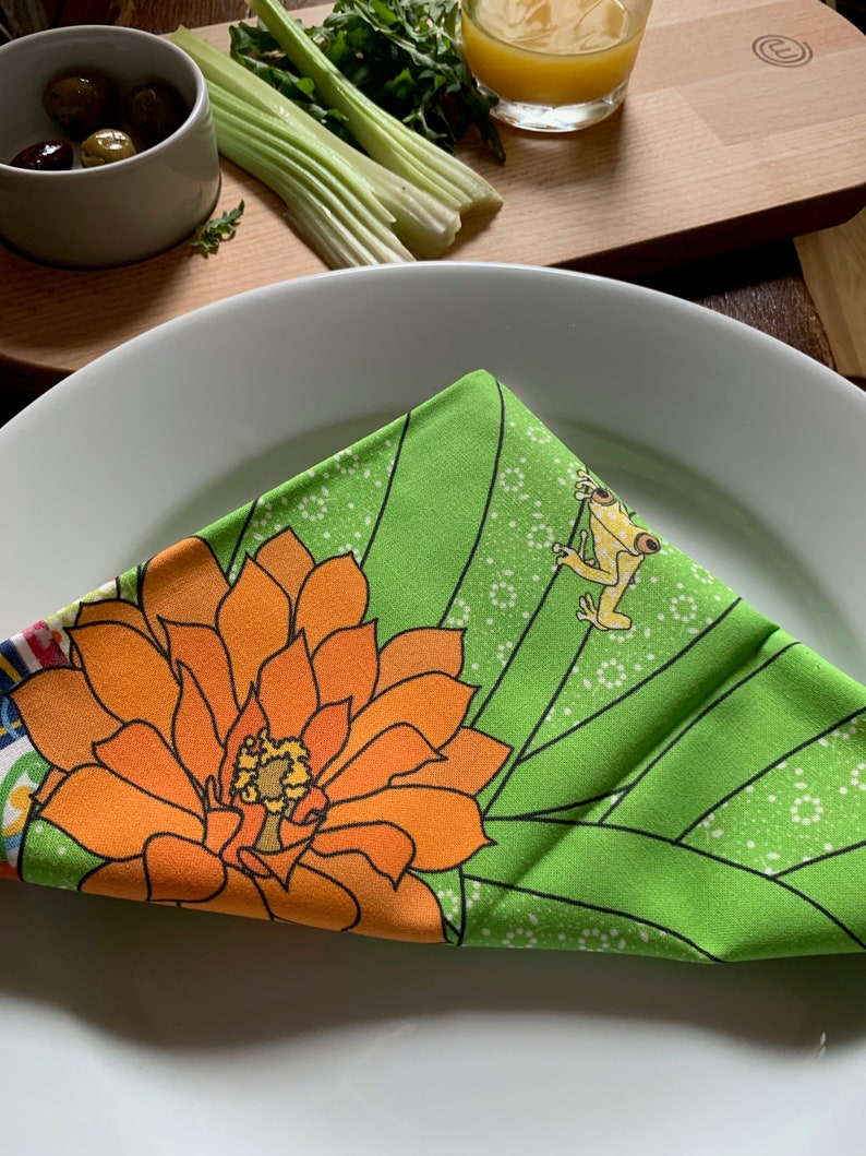 Puede incluir: Una servilleta plegada con estampado floral verde y naranja sobre un plato blanco. La servilleta presenta una gran flor naranja y una rana de dibujos animados. El fondo incluye una tabla de cortar de madera, apio, aceitunas y un vaso de zumo.