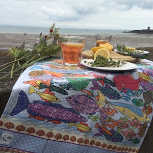 May include: A colorful tablecloth with a fish pattern, featuring a variety of fish in different colors and sizes. The tablecloth is laid out on a table with two glasses of orange juice and a plate of lemon slices and thyme.