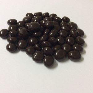 Puede incluir: Un montón de almendras cubiertas de chocolate negro. Las almendras son de forma ovalada y el chocolate es de color marrón oscuro.