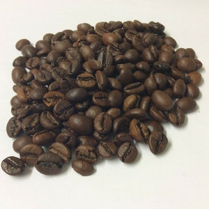 Puede incluir: Primer plano de un montón de granos de café marrones oscuros sobre un fondo blanco.