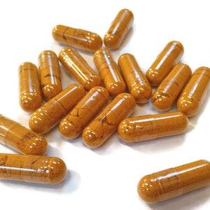 Op de afbeelding: Een close-up van een stapel oranje capsules op een wit oppervlak. De capsules zijn gevuld met een geel poeder.