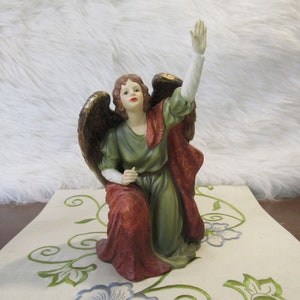May include: A kneeling angel figurine with outstretched arm, wearing a green robe and a red cloak with gold accents. The angel has brown hair and a white face.