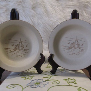 May include: Two beige ceramic bowls with a brown lighthouse design. The bowls are on black stands.