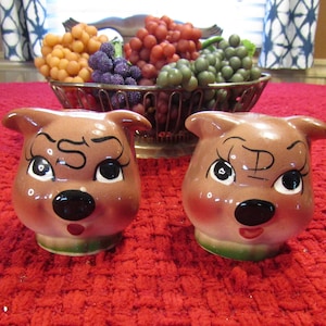May include: A pair of ceramic salt and pepper shakers shaped like pig faces. The shakers are brown with black eyes, noses, and mouths. The salt shaker has an "S" painted on its forehead, and the pepper shaker has a "P" painted on its forehead.