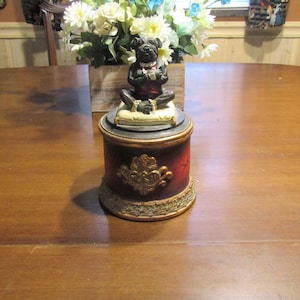 May include: A black ceramic dog figurine wearing a tuxedo sits on a cream-colored pillow atop a red and gold decorative jar with a gold floral design.
