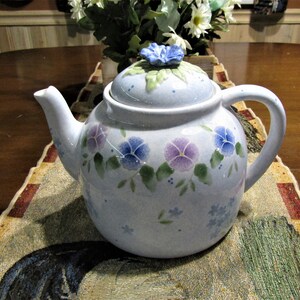 May include: A blue and white ceramic teapot with a floral design. The teapot has a lid with a blue flower on top. The teapot is sitting on a table with a floral tablecloth.