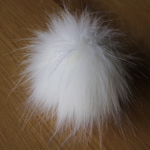 May include: A fluffy white pom pom made of faux fur. The pom pom is on a wooden surface.