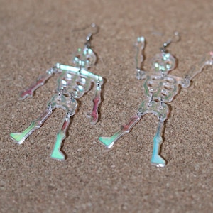 May include: A pair of iridescent skeleton earrings. The earrings are made of clear acrylic and have a holographic finish. The skeletons are posed in a playful, dancing position.