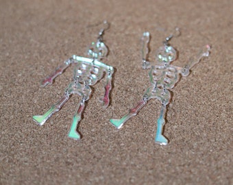 May include: A pair of iridescent skeleton earrings. The earrings are made of clear acrylic and have a holographic finish. The skeletons are posed in a playful, dancing position.