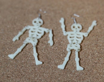 May include: A pair of skeleton earrings made of white plastic. The earrings are dangling and have a silver hook.
