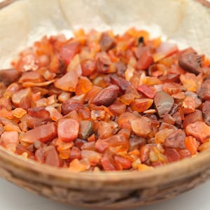 May include: A wooden bowl filled with small, polished carnelian gemstones. The stones are a mix of orange, red, and brown hues, with a translucent quality. The bowl's rim is a darker brown, contrasting with the bright stones. The gemstones are a popular choice for jewelry and decorative purposes.
