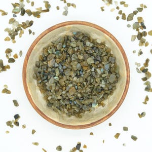 May include: A wooden bowl filled with small, irregularly shaped, green and brown gemstones. The bowl has a light brown rim and sits on a white surface. Scattered around the bowl are additional gemstones.