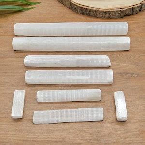 May include: A collection of seven white selenite crystal wands arranged in a tiered formation. The crystals are elongated, rectangular, and feature a fibrous, translucent texture with a natural, slightly reflective sheen across their surfaces.