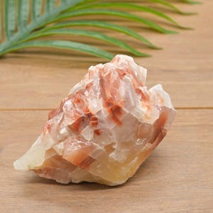 May include: A natural calcite crystal, displaying a mix of white, cream, and reddish-brown colours. The rough stone has a crystalline structure and is shown on a wooden surface, with a green leaf in the background.