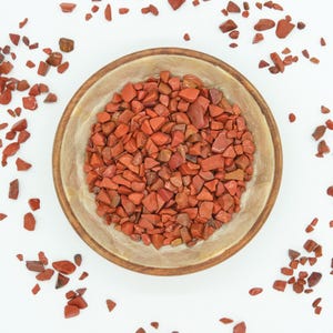 May include: A wooden bowl containing numerous small, irregularly shaped, reddish-brown jasper stones. The bowl has a light-coloured interior and a darker, wooden rim. Jasper pieces are scattered around the bowl on a white surface.