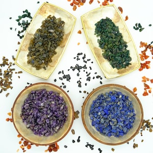 May include: Assortment of colourful gemstone chips in decorative bowls and on a white surface. The chips are in shades of green, purple, blue, and brown.