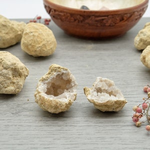 May include: Beige geodes, one split open to show a crystal interior. A carved wooden bowl and pink berries are in the background. The geodes are on a grey wooden surface.
