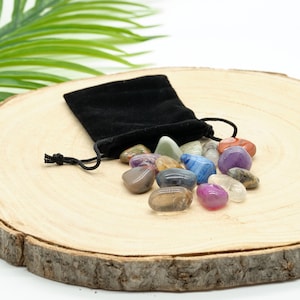 May include: A black velvet pouch with a drawstring closure, containing a collection of tumbled stones in various colours and shapes. The stones are arranged on a wooden surface.