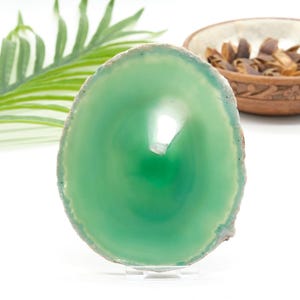 May include: A polished, oval green agate slice, displayed on a clear stand. The stone has concentric rings of varying shades of green, with a lighter centre. The outer edge has a rough, natural texture. Decorative item.
