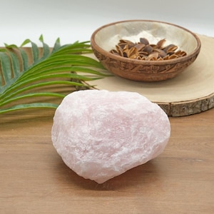 May include: A large, raw, pale pink rose quartz crystal with a rough, crystalline texture and irregular shape. The stone displays a translucent to opaque appearance with natural white inclusions throughout its solid, chunky, and unpolished surface.