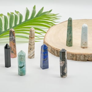 May include: A selection of gemstone obelisks in black, pink, blue, green, and brown. The obelisks are displayed on a white surface, with a wooden slice and green foliage in the background. Each obelisk is a unique shape.