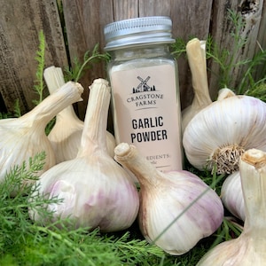 K&ouml;nnte beinhalten: Ein Glasgef&auml;&szlig; mit Knoblauchpulver und einem silbernen Deckel. Das Glas hat ein Etikett mit der Aufschrift "Cragstone Farms" und "Garlic Powder". Das Glas ist von mehreren ganzen Knoblauchknollen umgeben.