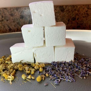 May include: A stack of six white marshmallows on a gray surface with dried lavender and chamomile flowers surrounding them.
