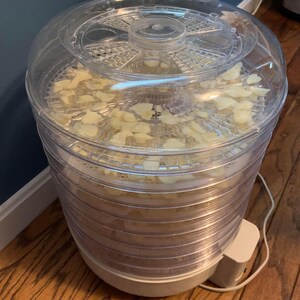 K&ouml;nnte beinhalten: Ein transparenter Lebensmitteldehydrator aus Kunststoff mit mehreren Tabletts, die mit Apfelscheiben gef&uuml;llt sind. Der Dehydrator hat einen wei&szlig;en Sockel und einen transparenten Deckel.