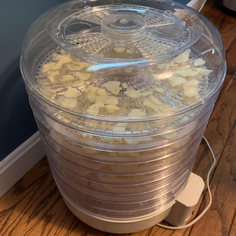 K&ouml;nnte beinhalten: Ein transparenter Lebensmitteldehydrator aus Kunststoff mit mehreren Tabletts, die mit Apfelscheiben gef&uuml;llt sind. Der Dehydrator hat einen wei&szlig;en Sockel und einen transparenten Deckel.