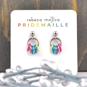 May include: A pair of silver Pridemaille earrings with a rainbow of coloured rings hanging from each earring. The earrings are displayed on a white card with the text "rebeca mojica PRIDEMAILLE" printed on it.