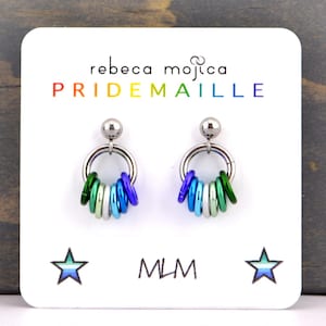 May include: A pair of silver hoop earrings with a rainbow of coloured rings hanging from each hoop. The earrings are on a white card with the text "rebeca mojica PRIDEMAILLE MLM" printed on it.