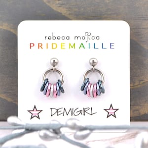 May include: A pair of silver earrings with a hoop design and a cluster of pink, blue, and grey rings hanging from each hoop. The earrings are displayed on a white card with the text "rebeca mojica PRIDEMAILLE DEMIGIRL" printed on it.