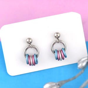 May include: A pair of silver stud earrings with a circle of blue and pink rings hanging from each. The earrings are displayed on a white card.