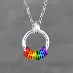 May include: A silver necklace featuring a large circular pendant. A series of small, colourful rings in red, orange, yellow, green, blue, and purple are attached to the bottom of the pendant, creating a rainbow effect.