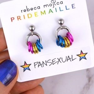 May include: A pair of silver earrings with a ring of colorful metal rings hanging from each. The rings are pink, blue, and yellow, representing the pansexual pride flag. The earrings are displayed on a white card with the text "rebeca mojica PRIDEMAILLE PANSEXUAL" printed on it.