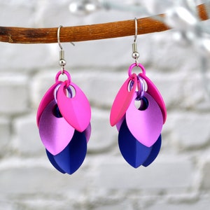May include: Handmade earrings with a scale design. The earrings feature pink, purple, and blue metallic scales, suspended from silver-coloured ear wires. The earrings are displayed against a blurred background, with a wooden branch.